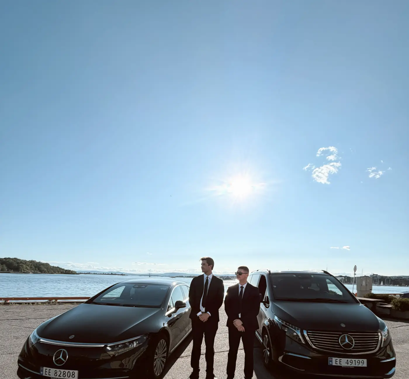 To Husby-sjåfører i dress mellom en Mercedes EQS og EQV ved Oslofjorden under klar himmel